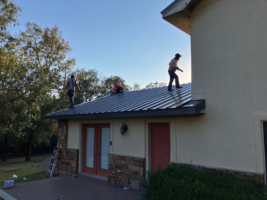 Workers installing a metal roof on a house under clear blue skies.