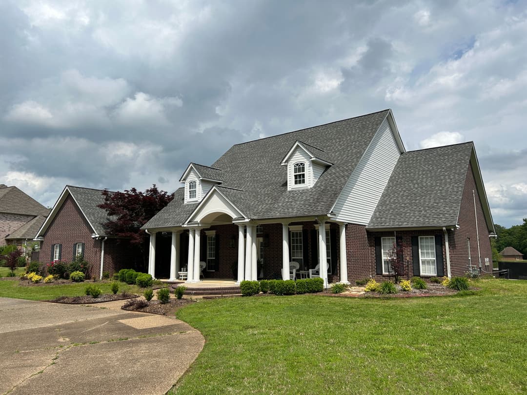 Large brick house with a gabled roof, white columns, and green lawn under a cloudy sky.