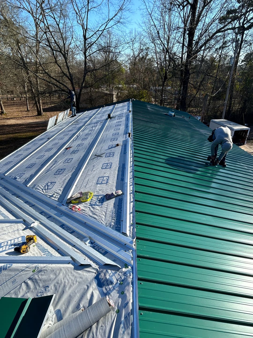 Workers installing green metal roofing on a residential home under a clear blue sky.