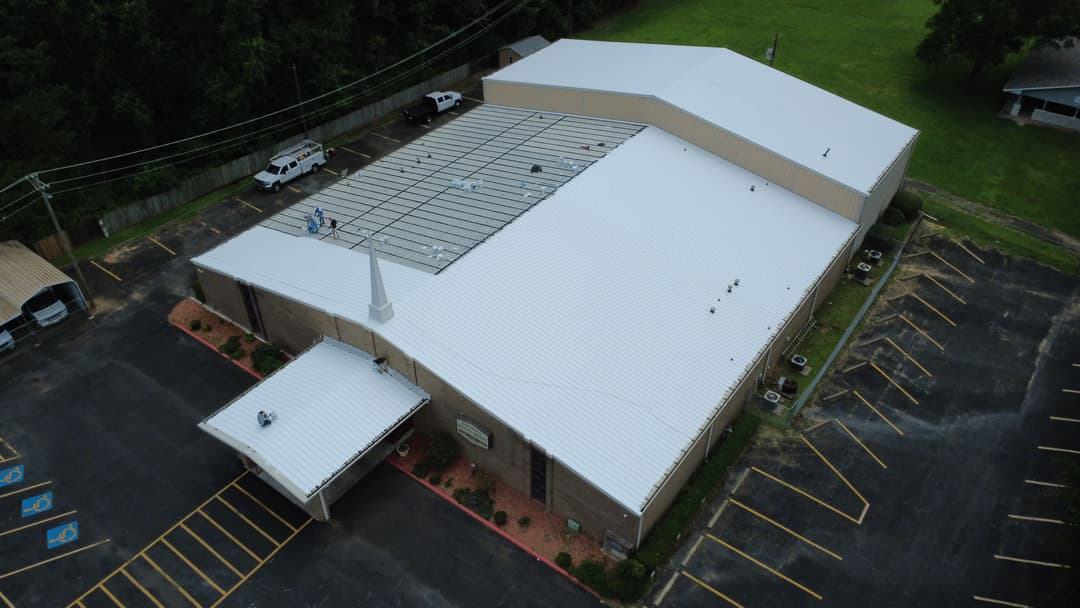 Aerial view of a large building with a white metal roof and paved parking lot.