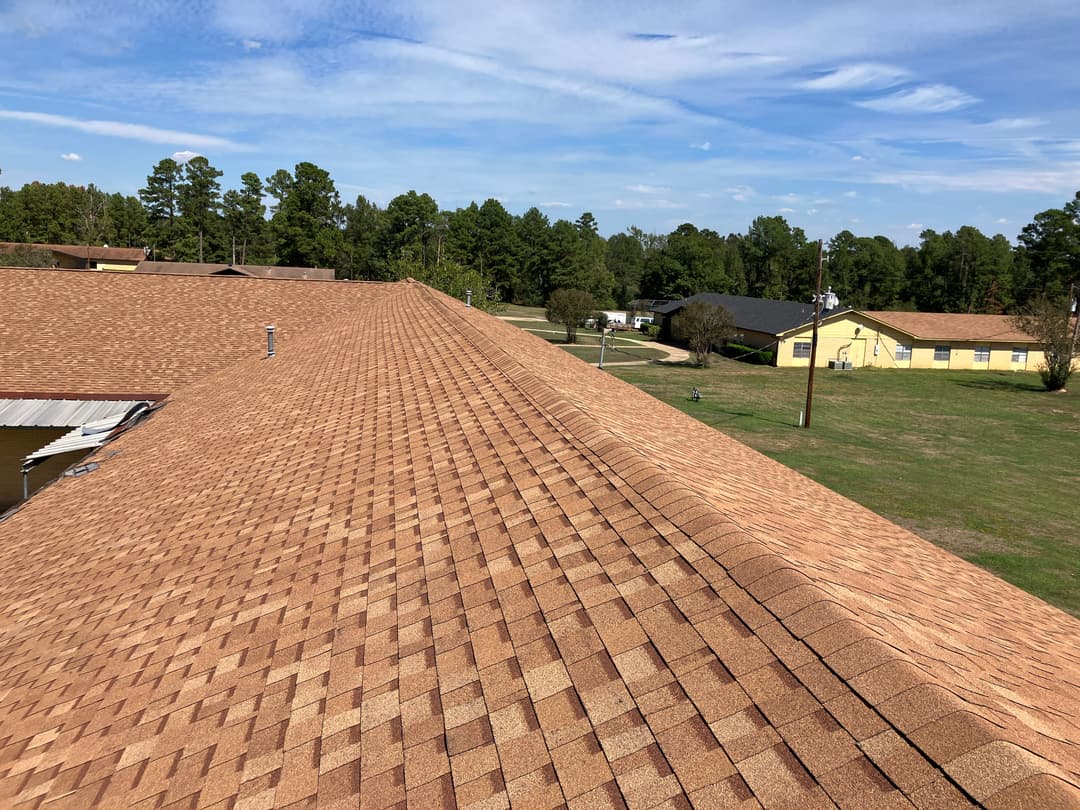 Aerial view of a brown shingle roof with trees and buildings in the background.
