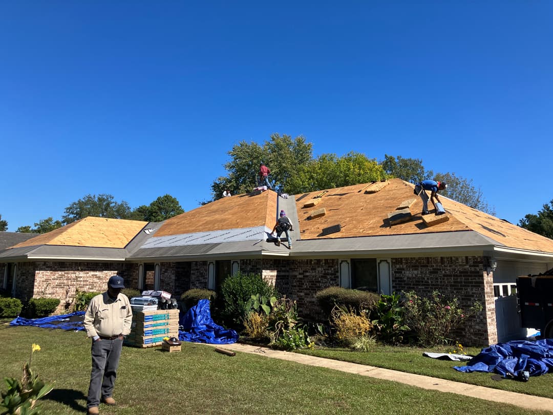 Roofing crew replacing shingles on a house with blue sky and greenery in the background.