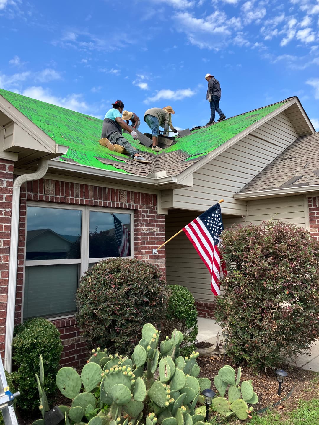 Roofers applying green coating on a house with an American flag and cacti in the foreground.