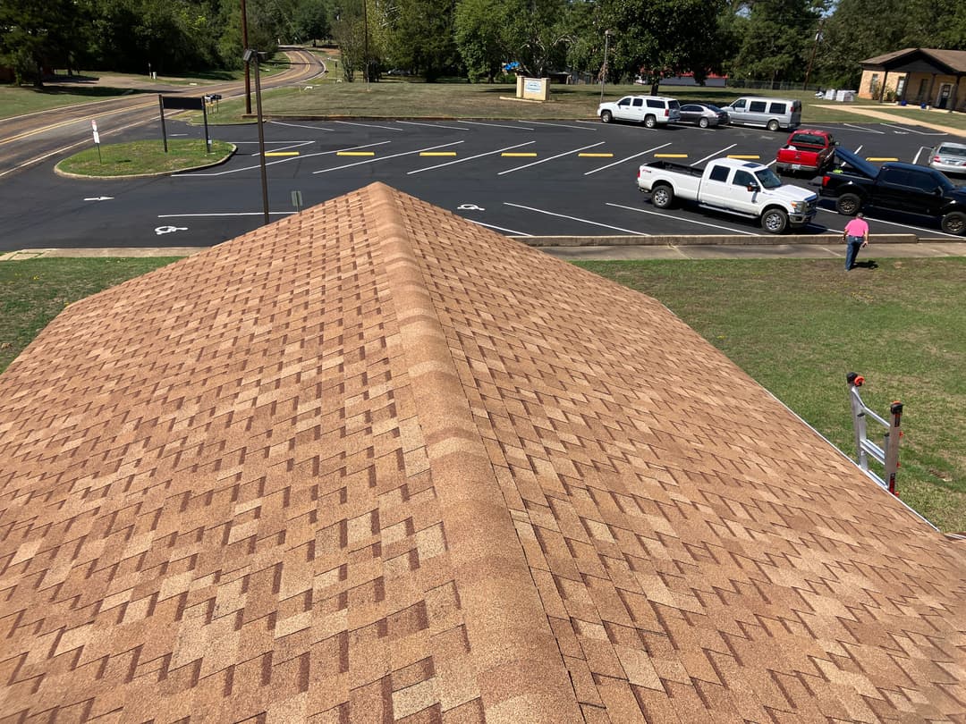 View of a brown shingle roof with a parking lot and trees in the background.