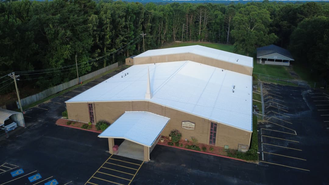 Aerial view of a church with a white roof, surrounded by trees and a parking lot.