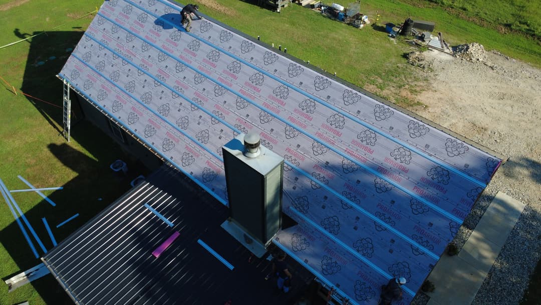 Workers installing roofing materials on a house with a chimney and open yard.